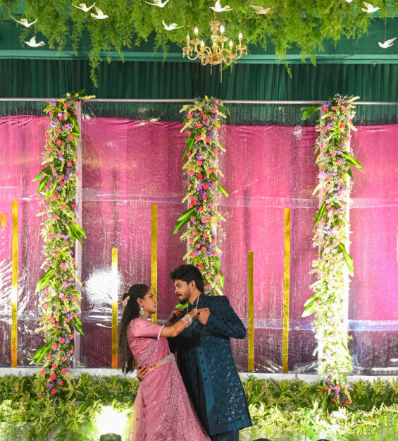 A couple in traditional attire dances on a decorated stage with pink and green floral arrangements and a leafy ceiling backdrop.